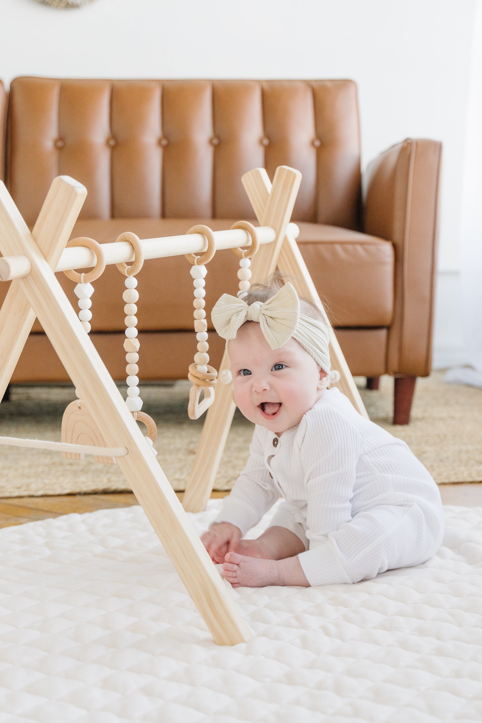 A baby play gym made of natural pine wood with a frame and four toys attached, displayed on the floor with a baby lying on it.