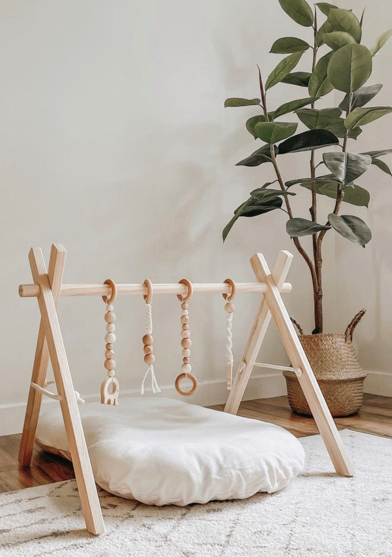 A baby play gym made of natural pine wood with a frame and four toys attached, displayed on the floor with a baby lying on it.