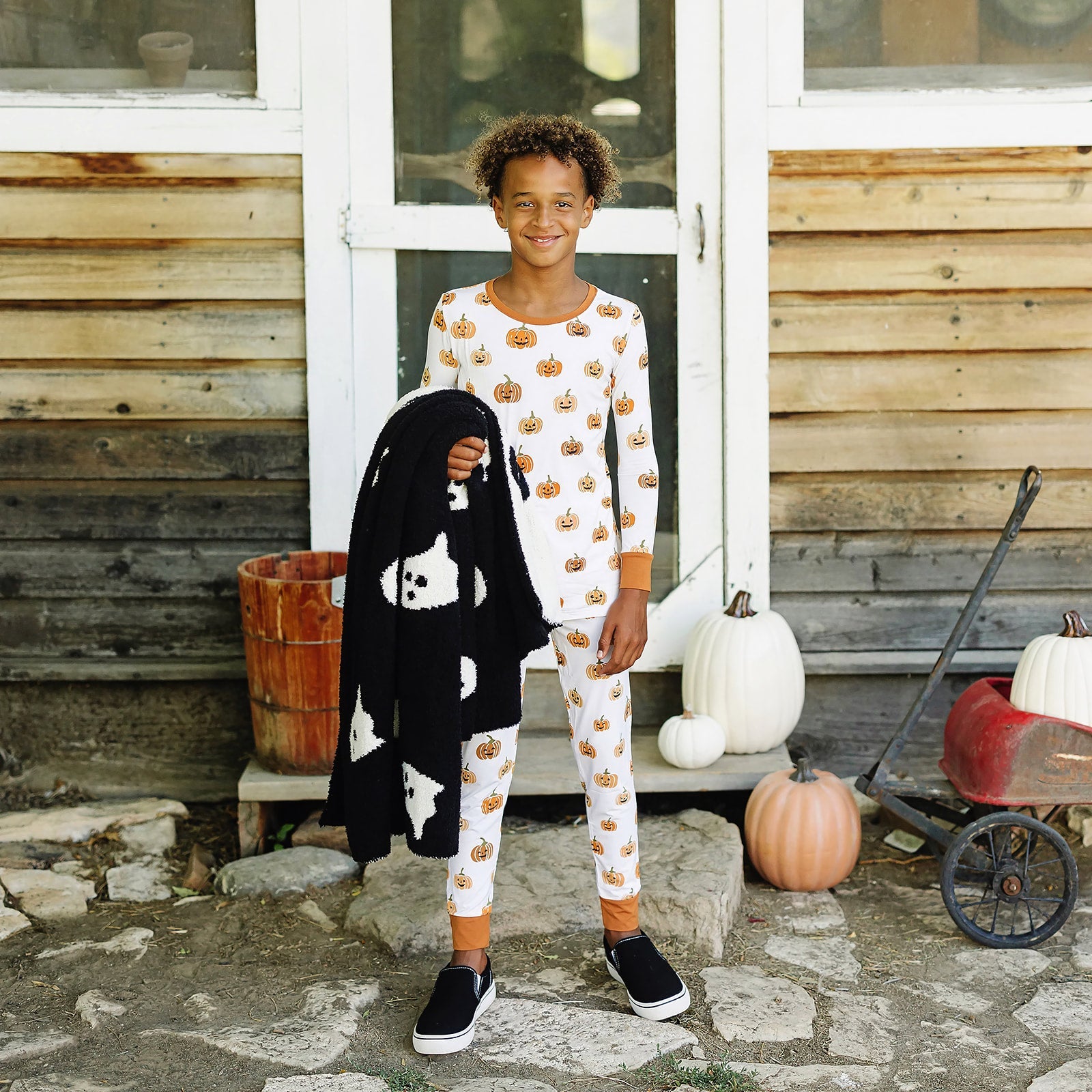 Child wearing a Halloween-themed outfit with pumpkins and ghosts, standing in front of a rustic wooden building.
