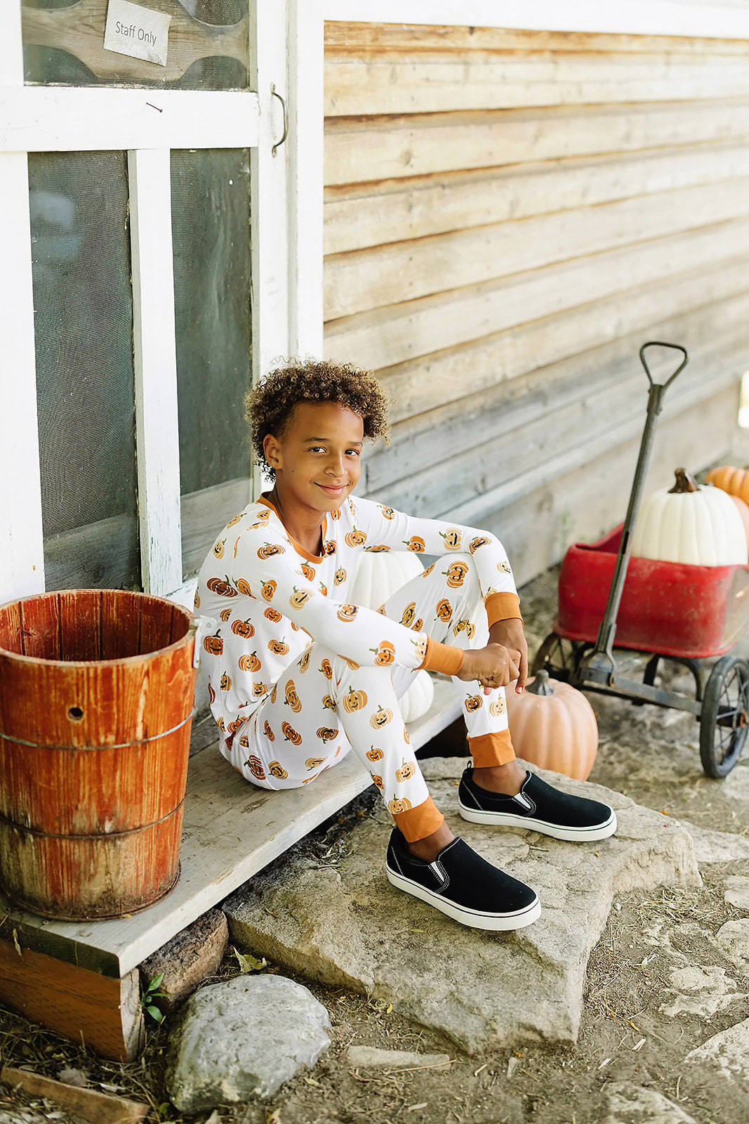 Child in Halloween pajamas sitting on a stone step with a wooden bucket and pumpkin nearby.