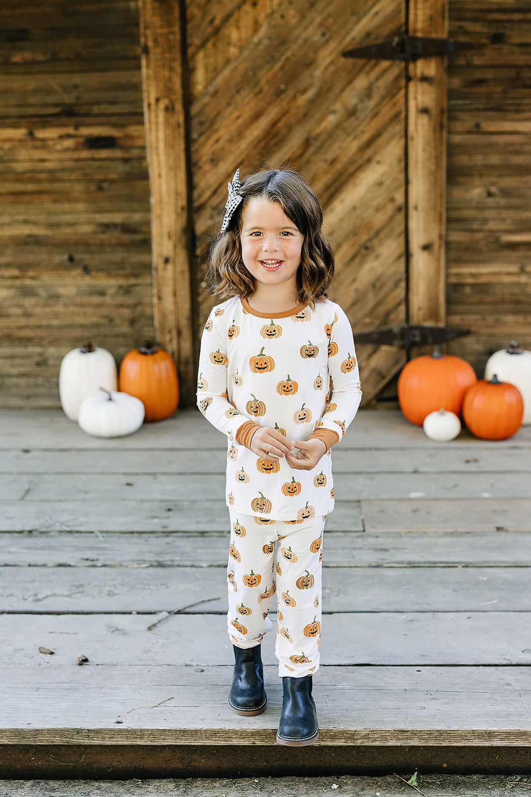 Child wearing a pumpkin-patterned outfit standing on a wooden deck with pumpkins in the background.