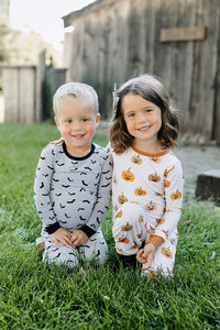 Two children in Halloween-themed pajamas sitting on grass in front of a wooden building.