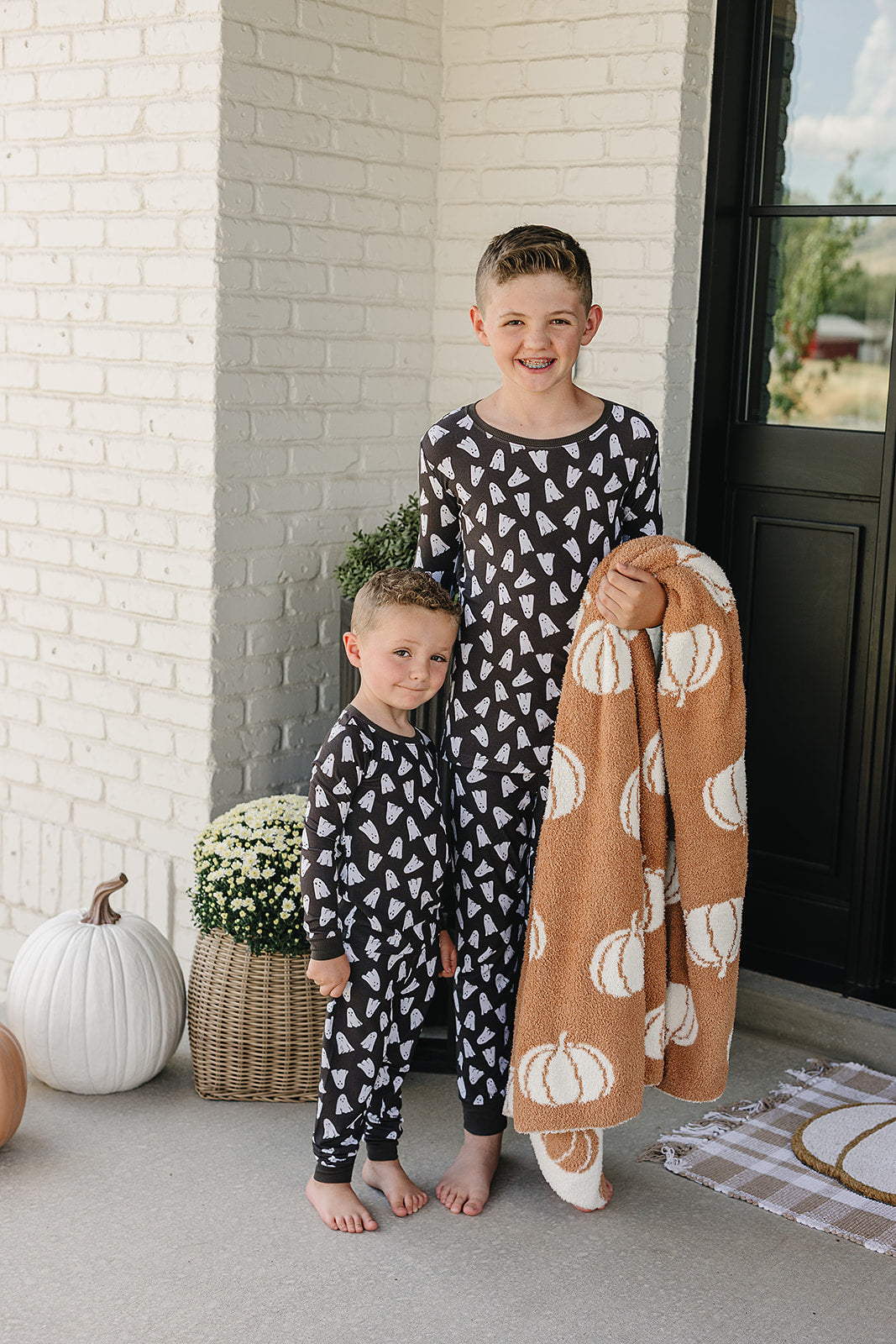 Two children in matching Halloween pajamas standing on a porch with a blanket and pumpkins.