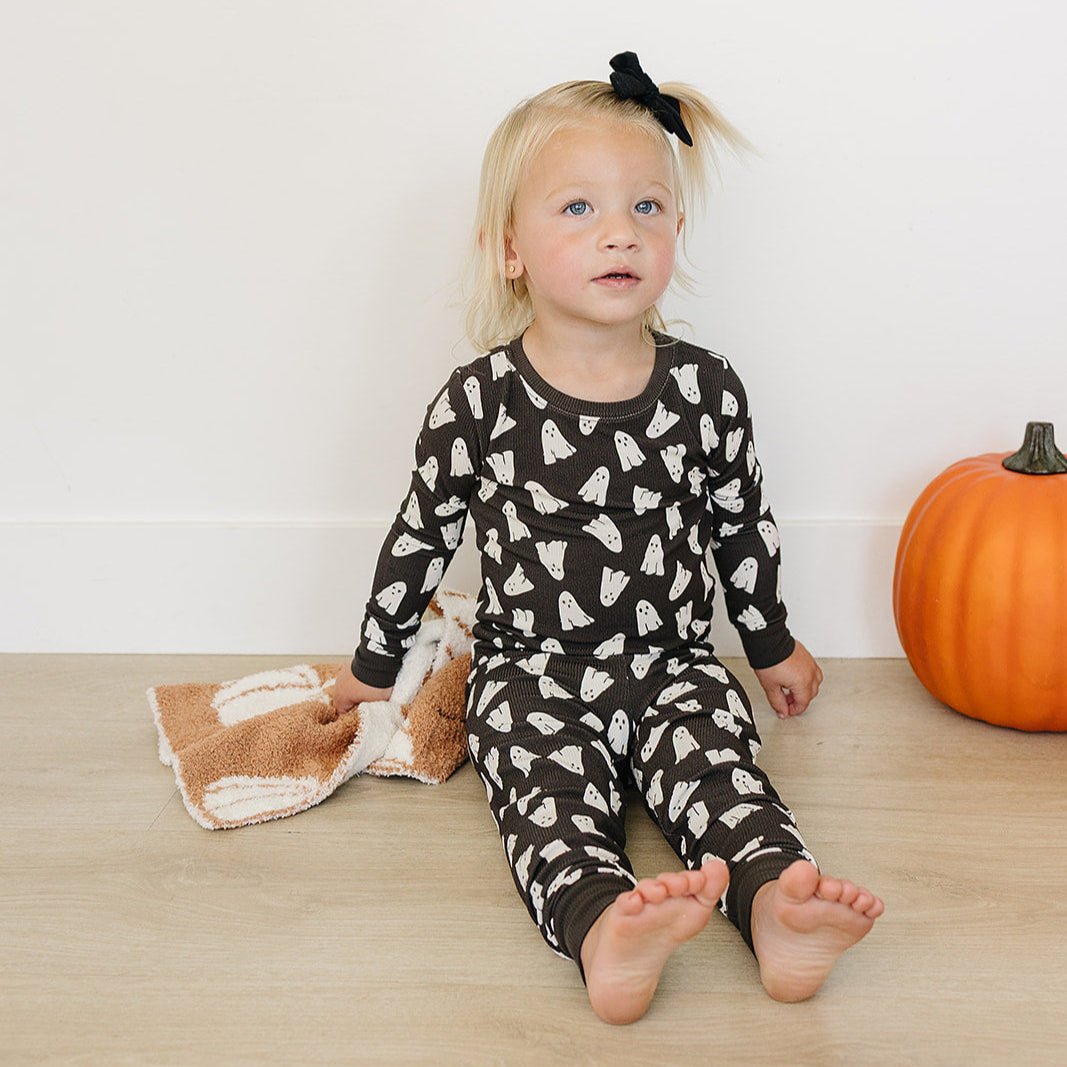 Child wearing a black patterned Halloween pajamas sitting on a wooden floor with a pumpkin in the background.
