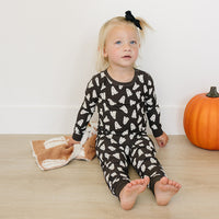 Child wearing a black patterned Halloween pajamas sitting on a wooden floor with a pumpkin in the background.