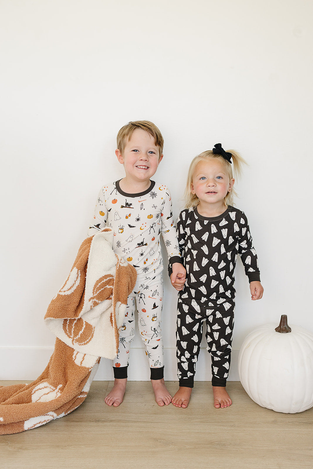 Two children in Halloween pajamas standing next to a white pumpkin on a wooden floor.