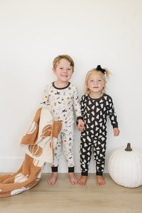 Two children in Halloween pajamas standing next to a white pumpkin on a wooden floor.