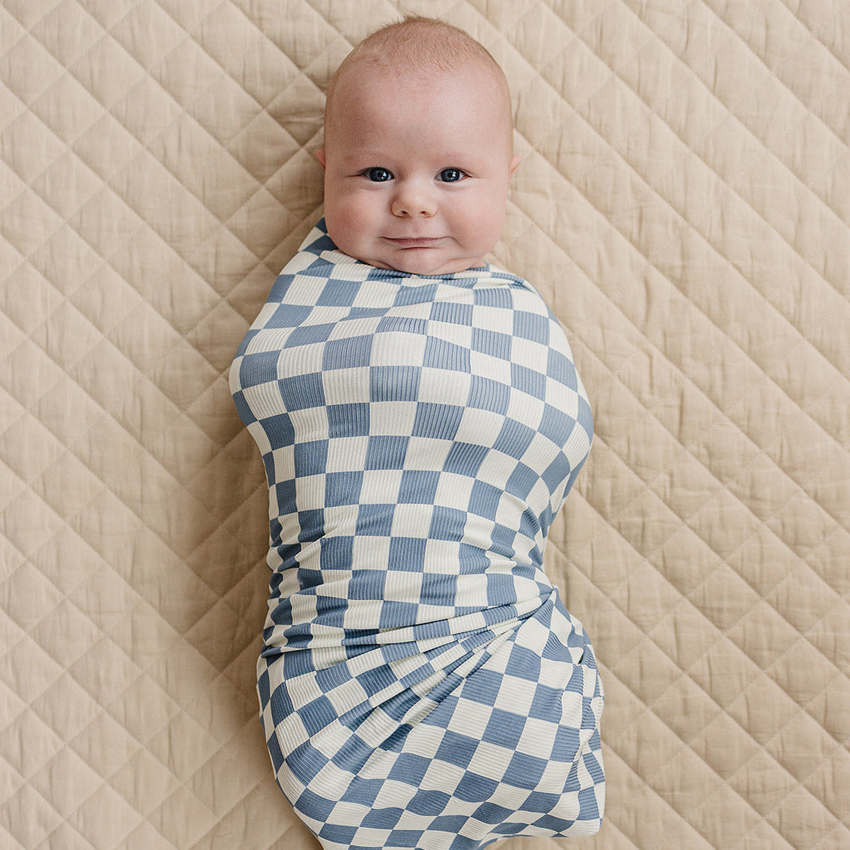 Baby swaddled in a blue and white checkered blanket on a textured beige surface