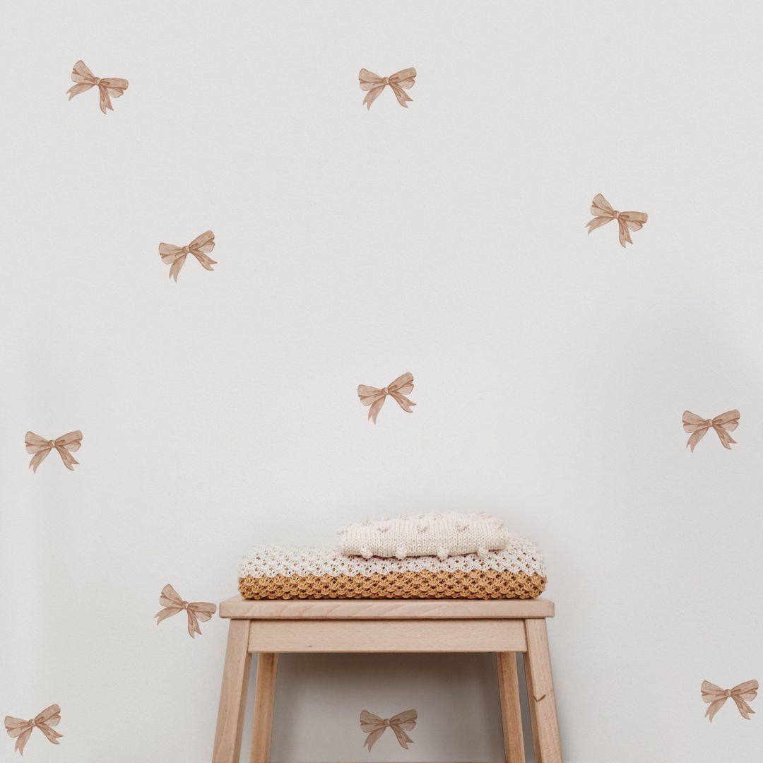 A set of beige bow decorating stickers for walls arranged on a white wall, with a wooden stool and a basket of blankets placed in front.