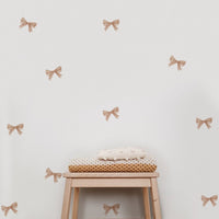 A set of beige bow decorating stickers for walls arranged on a white wall, with a wooden stool and a basket of blankets placed in front.